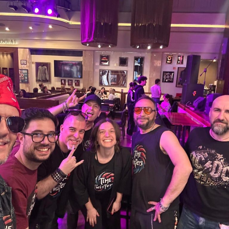 Un gruppo di sei persone sorridenti in un locale, indossando occhiali da sole e abbigliamento rock.