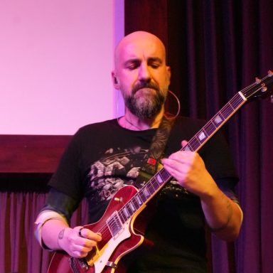 Un musicista con chitarra elettrica suona sul palco, vestito con una maglietta scura.